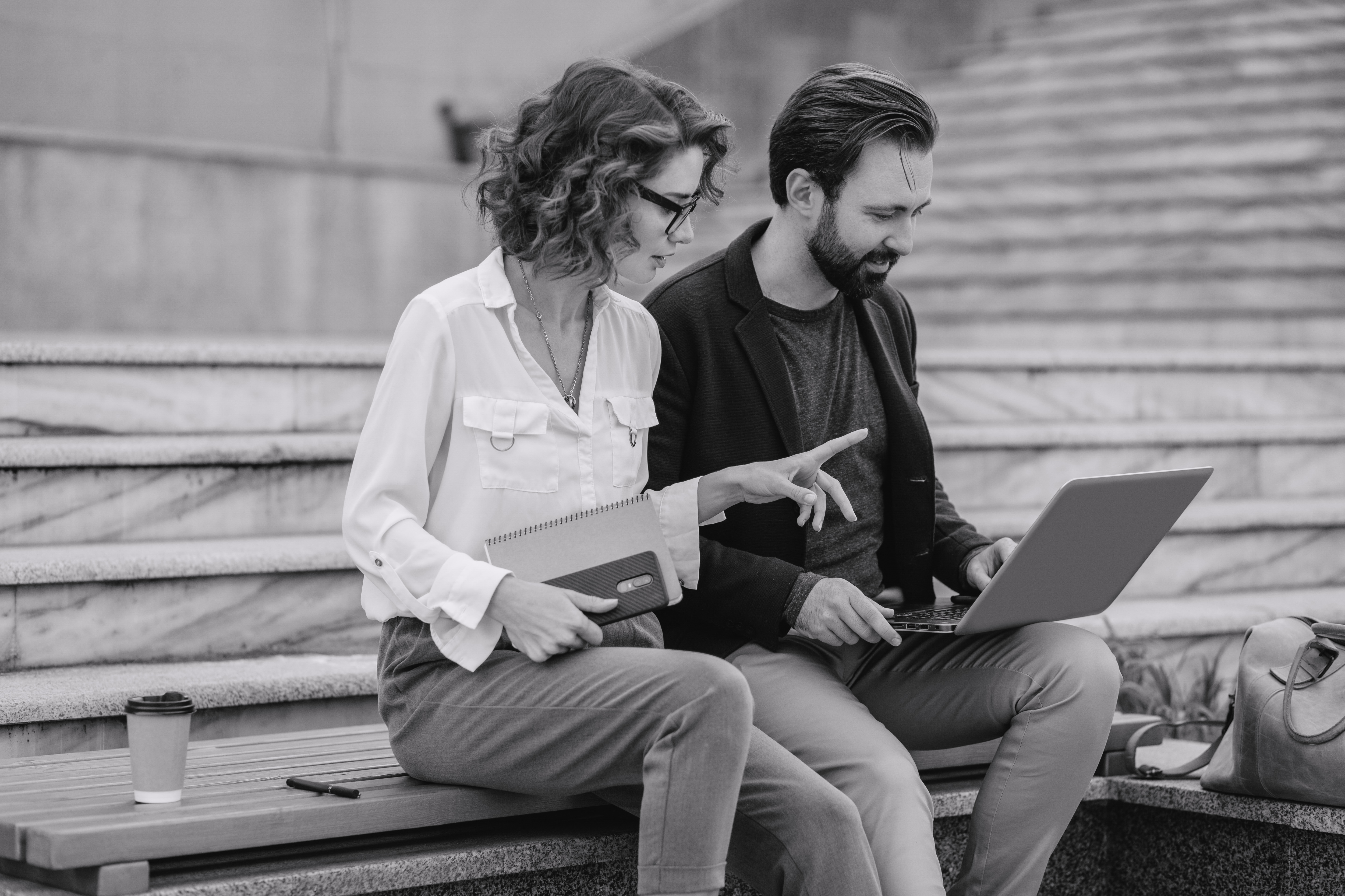 Ein Mann und eine Frau sitzen nebeneinander auf einer Treppe im städtischen Zentrum. Sie wirken wie ein attraktives Paar, das entspannt miteinander spricht. Vor sich haben sie einen Laptop, an dem sie gemeinsam arbeiten. Die urbane Umgebung im Hintergrund deutet auf eine moderne Innenstadt hin.
