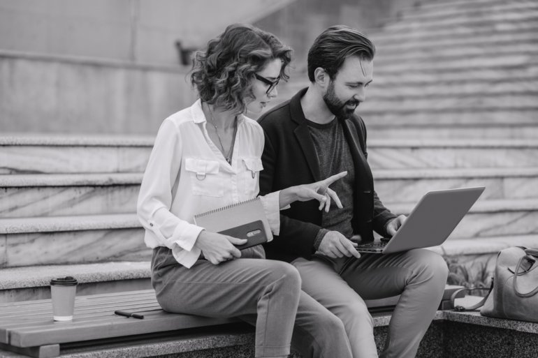 Ein Mann und eine Frau sitzen nebeneinander auf einer Treppe im städtischen Zentrum. Sie wirken wie ein attraktives Paar, das entspannt miteinander spricht. Vor sich haben sie einen Laptop, an dem sie gemeinsam arbeiten. Die urbane Umgebung im Hintergrund deutet auf eine moderne Innenstadt hin.